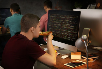 Young programmer eating pizza while working in office