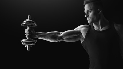 Black and white photo of handsome power athletic man in training pumping up muscles holding dumbbell stretching out his hand. Strong bodybuilder with perfect deltoid muscles, biceps, triceps