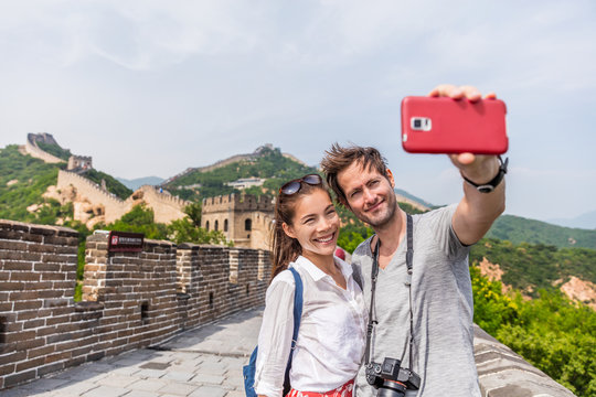 Happy couple tourists taking selfie picture at Great wall of china, top worldwide tourist destination. Young multiracial people using phone photography app for photos.