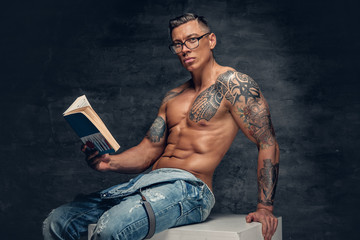 Shirtless muscular male with tattoo on a chest sits on a white wooden box and reads a book.