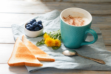 Breakfast idea. Cup with coffee with blueberries, dandelions and toast on linen napkin and wooden background