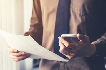 Young business man holding paper and using smartphone in the office with sunlight. Vintage toned filter.
