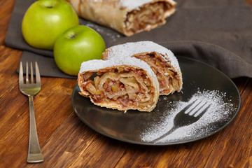 Apple strudel with icing sugar on black plate, wooden background