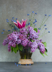Colorful bouquet in glass vase with purple lilac, pink tulip and blue forget-me-not flowers on grey textured background