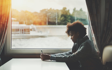 Beautiful pensive black Brazilian girl using smartphone while travelling on ship, thoughtful female teenager with curly afro hair holding cell telephone while sitting near window of ship cabin