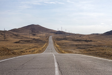 An empty curved cracked asphalt road to Lake Baikal is among the mountains in spring with clear sky and dry grass.