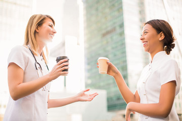 Two happy young nurses stand with cups of coffee and smile in time of a break, outdoors