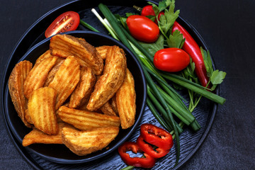 Fried potatoes on a black wooden plate with fresh vegetables - onions, tomatoes, parsley. Black background. Rustic