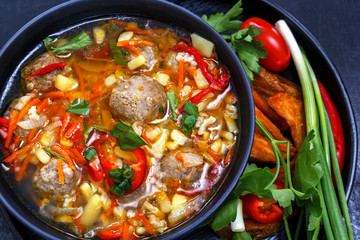 Soup with meatballs in a black plate, sliced vegetables - onion, parsley, pepper, tomato on a black background