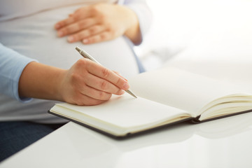 Young pregnant woman writes to her diary on a white table