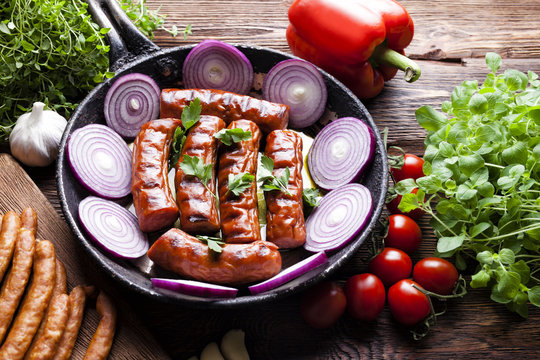 Grilled sausage with onion in a pan, raw meat and vegetables on wooden table