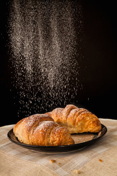 Two fresh croissants being dusted with icing sugar