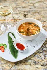 Korean cuisine. Spicy kimchi soup in white ceramic bowl served with red sauce in spoon, green leaf and paprika rings with glass of white wine on marble table.