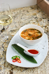 Korean cuisine. Spicy kimchi soup in white ceramic bowl served with red sauce in spoon, green leaf and paprika rings with glass of white wine on marble table.