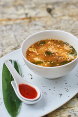 Korean cuisine. Spicy kimchi soup in white ceramic bowl served with red sauce in spoon, green leaf and paprika rings with glass of white wine on marble table.