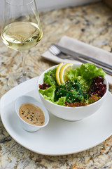 Seeweed salad in white ceramic bowl served with lemon slices, sauce, glass of white wine, fork and knife on marble table