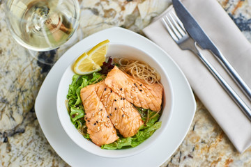 Asian cuisine. Warm salad with salmon fish and rice served in white ceramic bowl with knife, fork and glass of white wine on marble background. Flat lay or top view.