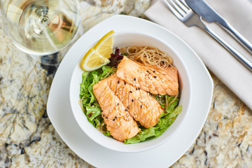Asian cuisine. Warm salad with salmon fish and rice served in white ceramic bowl with knife, fork and glass of white wine on marble background. Flat lay or top view.