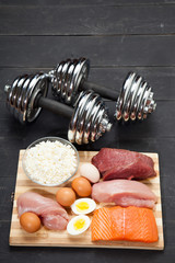 Proteins and dumbbells. Fish, cheese, eggs, meat, chicken  heart on a black wooden background