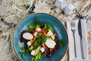 Greek salad with giant olives and provencal herbs served in blue ceramic plate with glass of white wine on marble table. Top view or flat lay.