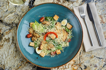 Cesar salad with baked chiken breast served in blue ceramic plate with glass of white wine, fork and spoon on textile napkin on marble table. Top view or flat lay.