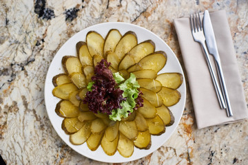Slices ofmarinated cucumber on white plate. Knife and fork on textile napkin on marble table. Top view or flat lay.