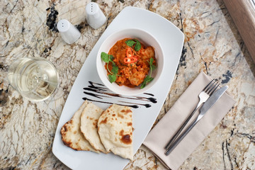 Chicken fillet slices fried in spicy cream sauce with tortilla bread served in white bowl with glass of wine, knife and fork on napkin on marble table
