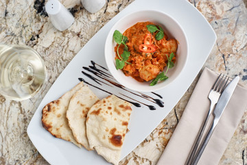 Chicken fillet slices fried in spicy cream sauce with tortilla bread served in white bowl with glass of wine, knife and fork on napkin on marble table