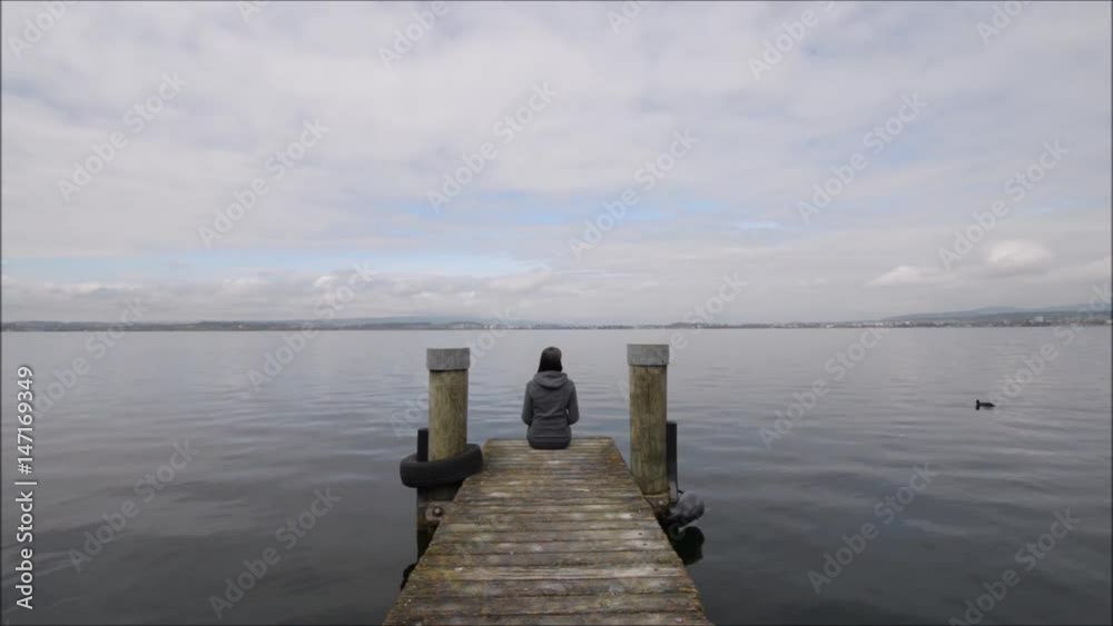 Wall mural a woman sits on a wooden pier on the lake shore. a duck is floating by. little attention to passing 