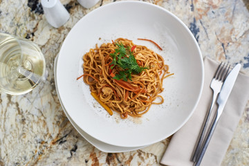 Flat egg noodles with vegetables served in big white ceramic round plate with glass of white wine on marble table