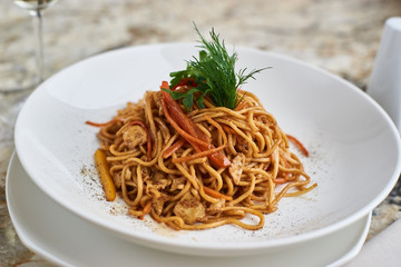 Flat egg noodles with vegetables served in big white ceramic round plate with glass of white wine on marble table