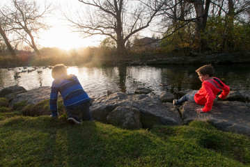 Two Young Caucasian Boy Brothers, Photographed from Behind, Play next to a Small Stream at Dusk