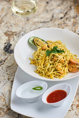 Flat egg noodles with seafood served in big white ceramic round plate with glass of white wine on marble table