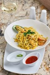 Flat egg noodles with seafood served in big white ceramic round plate with glass of white wine on marble table