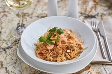 Rice noodles with peanuts, chicken and shrimps served on white round plate with glass of white white on marble table