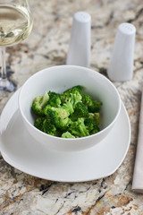 Steamed broccoli served in white bowl with glass of white wine on marble background