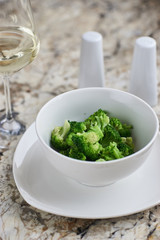 Steamed broccoli served in white bowl with glass of white wine on marble background