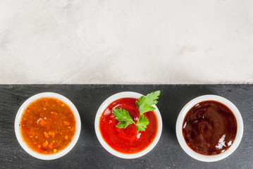 Three kinds of red tomato sauce on a white stone table: traditional classic ketchup, barbecue sauce, sweet and sour Chinese sauce. Top view copy space