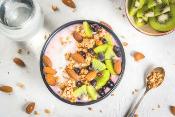 Healthy diet breakfast: smoothies bowl, with yogurt, fresh blueberries (black currant), kiwi, granola oatmeal, almond nuts. On a white stone table, with a glass of water and ingredients. Top view