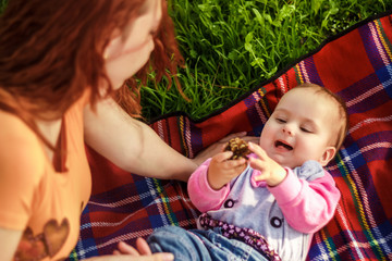 young beautiful woman playing with her baby on the plaid on green meadow