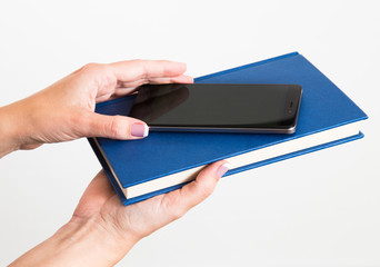 Closeup of young caucasian female hands holding modern smartphone and blue book isolated on white background. Choice concept. Horizontal color photo.