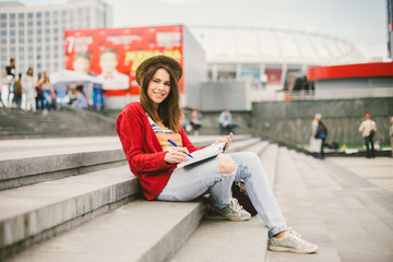 A beautiful, young Caucasian girl sitting on the street smile, the joy, sit with notebook and pen in Ruhi. In the red sweater, jeans and brown leather bag