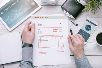 Man holds invoice of electric power usage over desk with tablet and smartphone with applications...