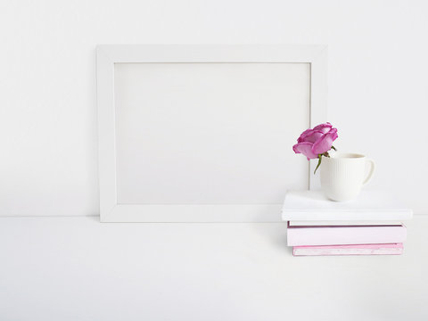 White blank wooden frame mockup with a rose flower in a porcelain cup and pile of books lying on the table. Poster product design. Styled stock feminine photography. Home decor.