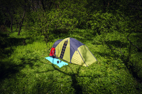 Tent in a green forest, red backpack, sticks, tourist boots, solar battery and tablet in sunny spring day