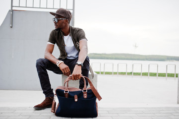 Portrait of sitting stylish african american man wear on sunglasses and cap outdoor. Street fashion black man.