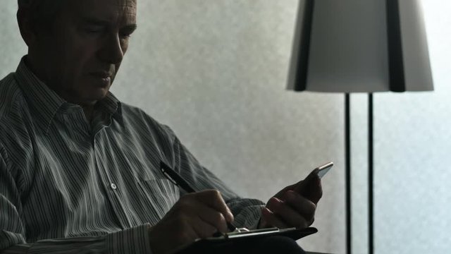 Elderly Man Using The Mobile Phone On The Couch And Writing On Notepad