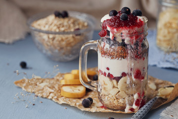 Layered dessert with muesli, yogurt, banana, linen seeds, sliced almonds and berries in glass mason jar on blue rustic background.