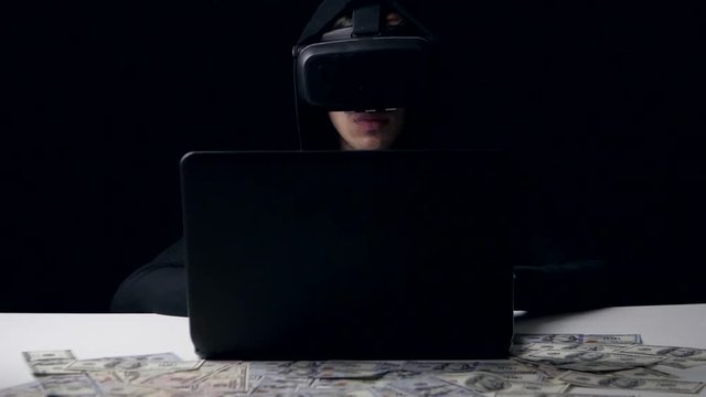 Hacker in the helmet of virtual reality work in dark room. He counts money. Cyber crime.