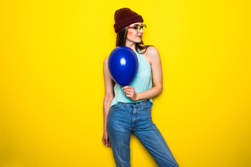 Cheerful hipster girl in red hat holding balloon on yellow wall